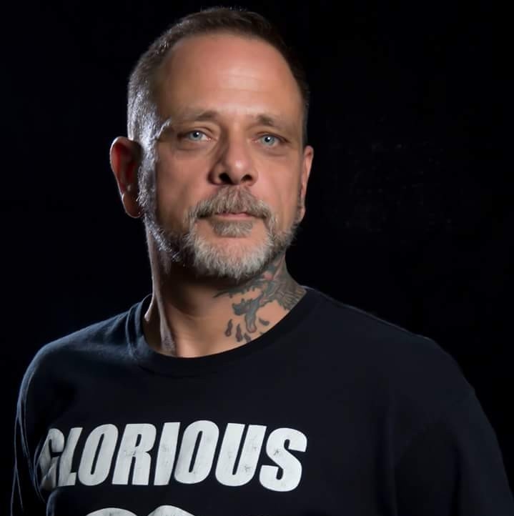 Portrait of a man with blue eyes and a gray stubble beard wearing a black T-shirt that says GLORIOUS, against a dark background, with a neck tattoo visible.