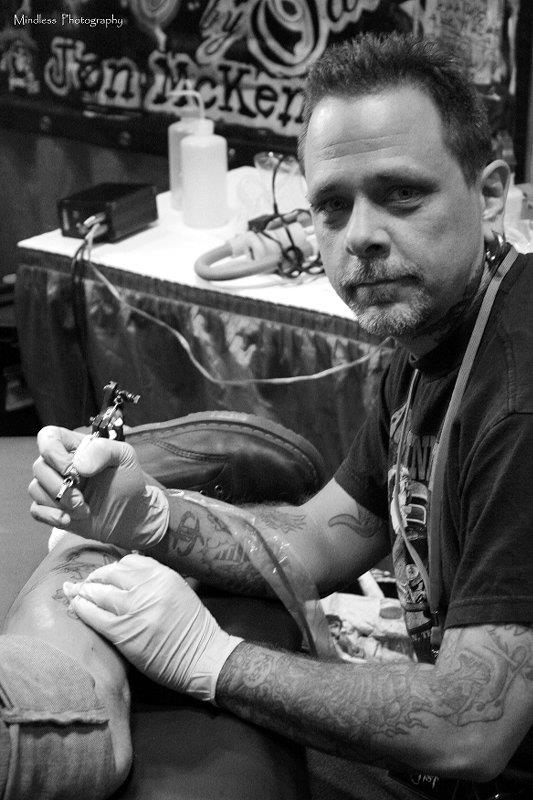 Tattoo artist wearing gloves, using a tattoo machine on a client’s leg in a studio setting (black and white).