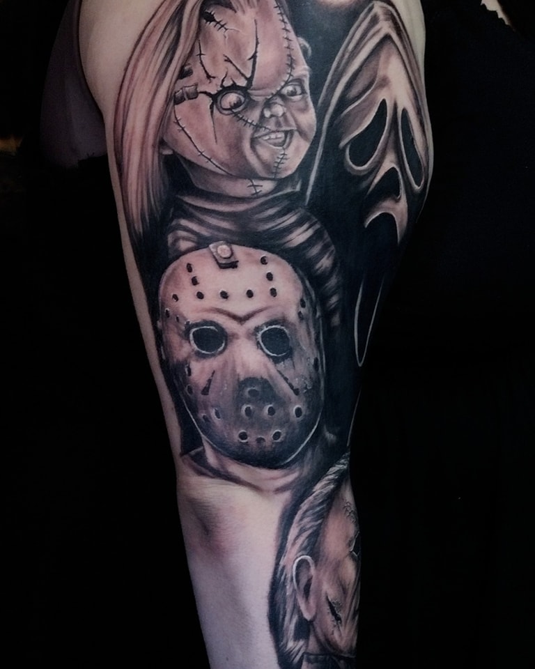 Sleeve tattoo showing a stitched-doll face (Chucky) above a Jason Voorhees hockey mask and a Ghostface-style mask on dark skin.