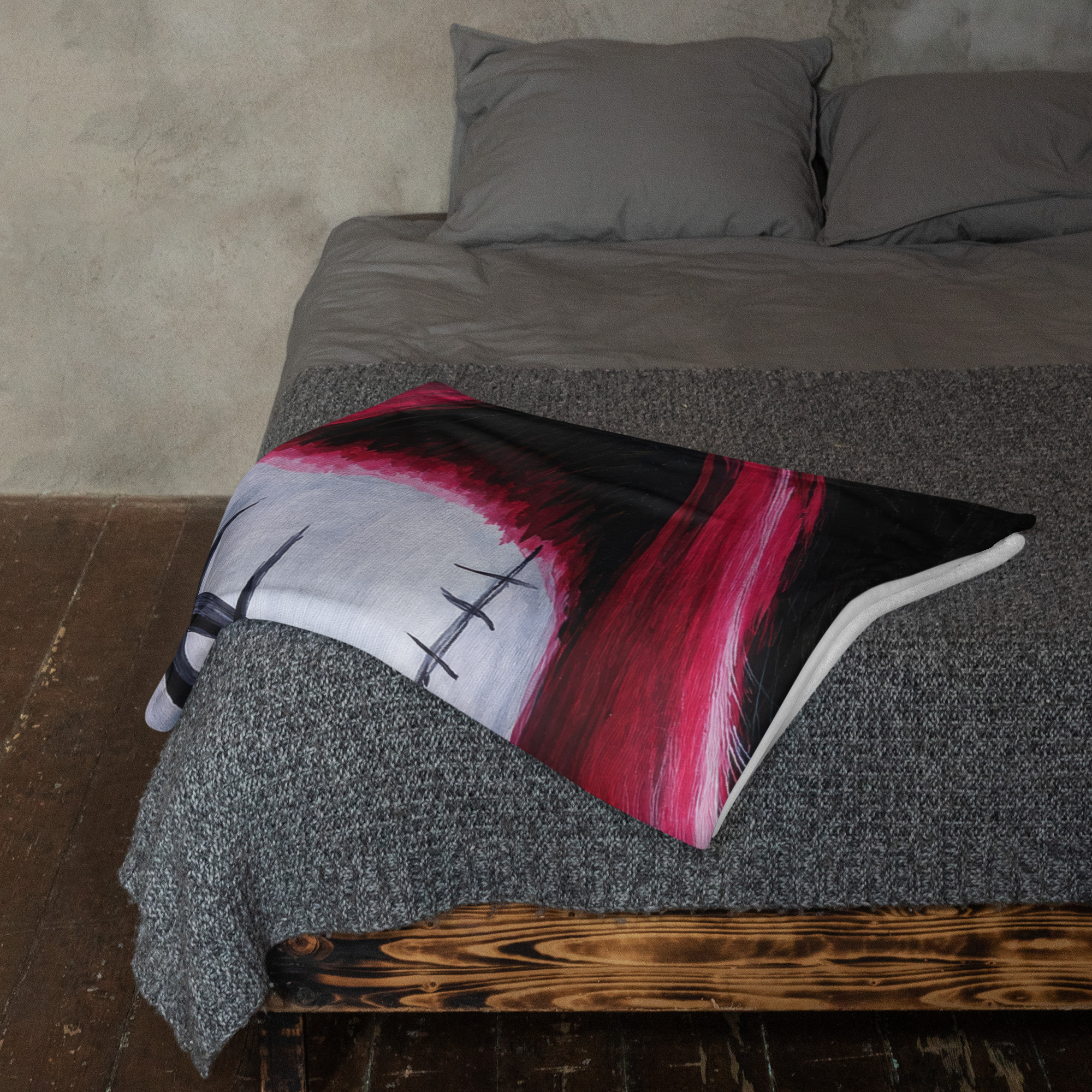 Top-down view of a bed with gray sheets and a bold red-and-black pillow on a dark rug and wooden bed frame visible at the foot of the bed.